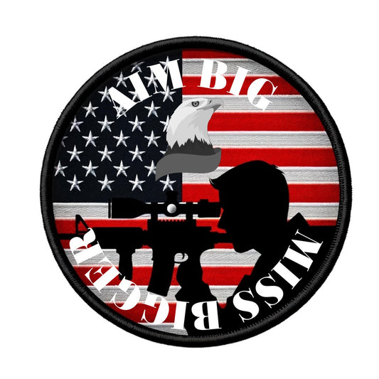 A circular military patch featuring a distressed American flag as the background. A silhouette of a figure aiming a rifle is centered on the patch, set against the stripes of the flag. Above and below the silhouette, in bold, stenciled lettering following the patch's curve, are the phrases "AIM BIG" and "MISS BIGGER", respectively. The overall design carries a satirical tone, suggesting a humorous take on military marksmanship.